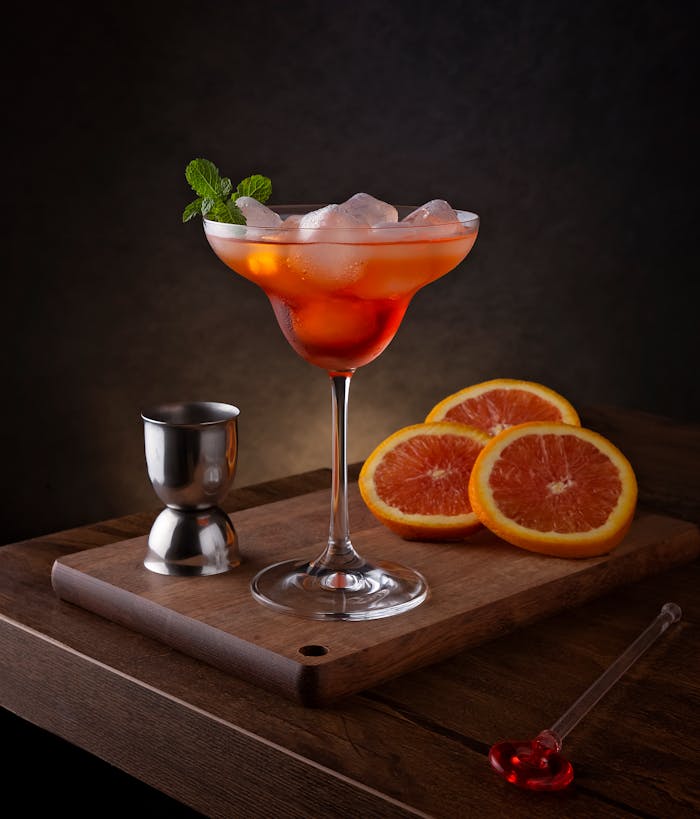 Close-up of an orange cocktail with ice and mint, surrounded by sliced oranges and bar tools.